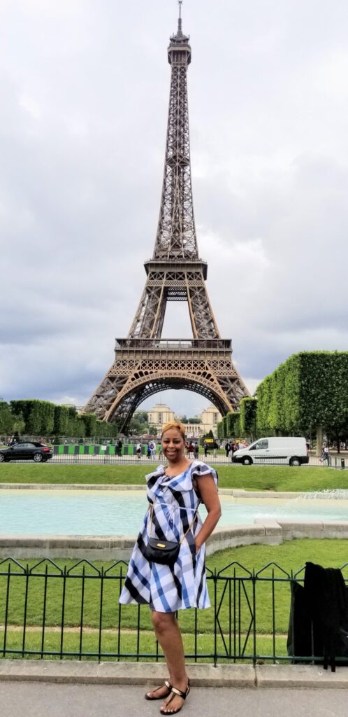 Tourist woman posing in front of the Eiffel Tower in Paris, wearing a blue and white patterned dress and sandals near a fountain.