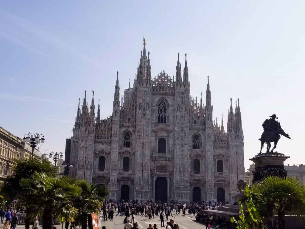 Front facade of a large Gothic cathedral with numerous spires and a crowd gathered in the square in front.