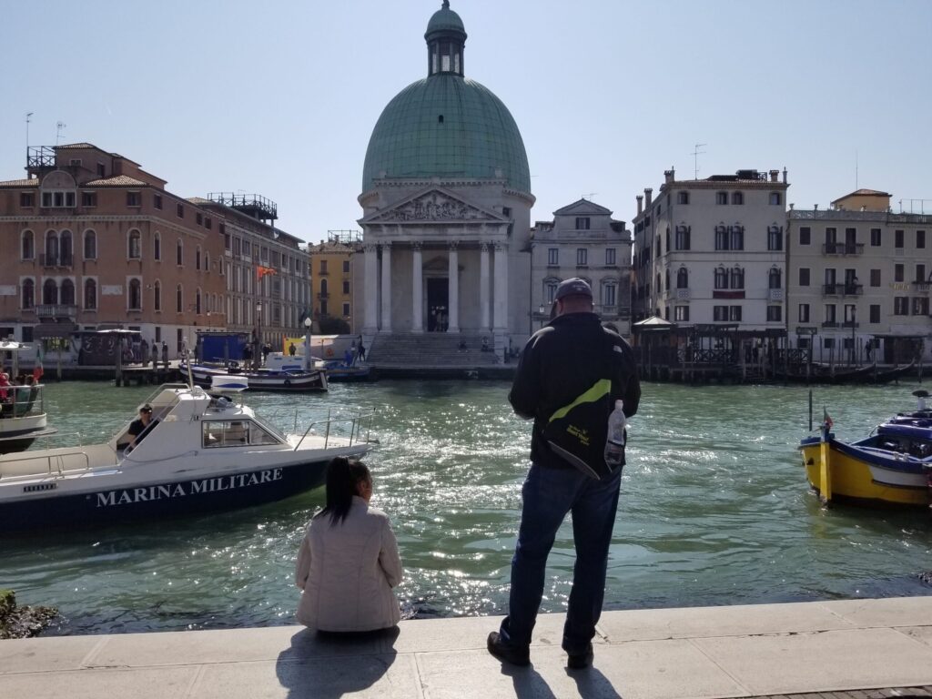 Two people stand by a canal in Venice, facing a large green-domed building with historic columns and pastel city buildings along the water; boats docked nearby.
