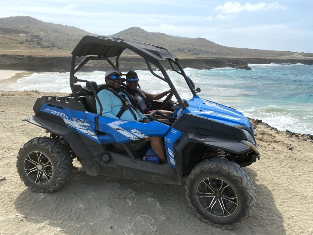 Two people sit in a blue off-road buggy on a sandy beach with blue ocean waves in the background, smiling at the camera.