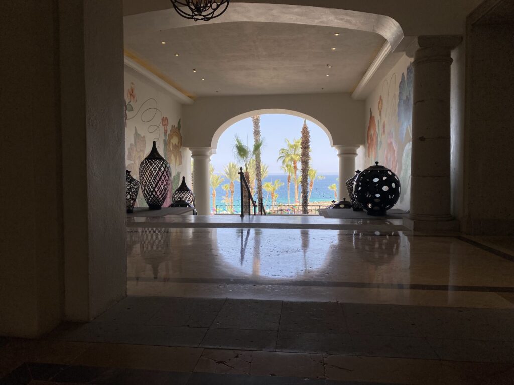 A stone colonnaded hallway opening to an outdoor seaside view with palm trees, seen through an arched doorway; decorative lanterns sit on a table in the foreground.