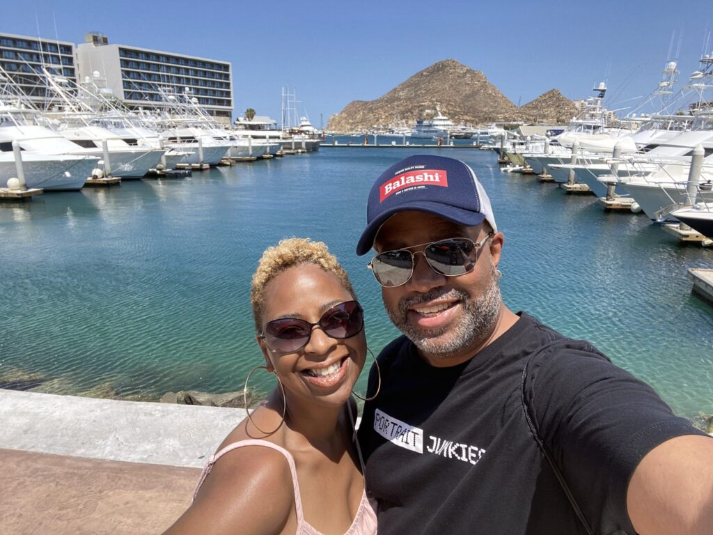 Smiling couple taking a selfie at a marina with white yachts, blue water, and rocky hills in the background.