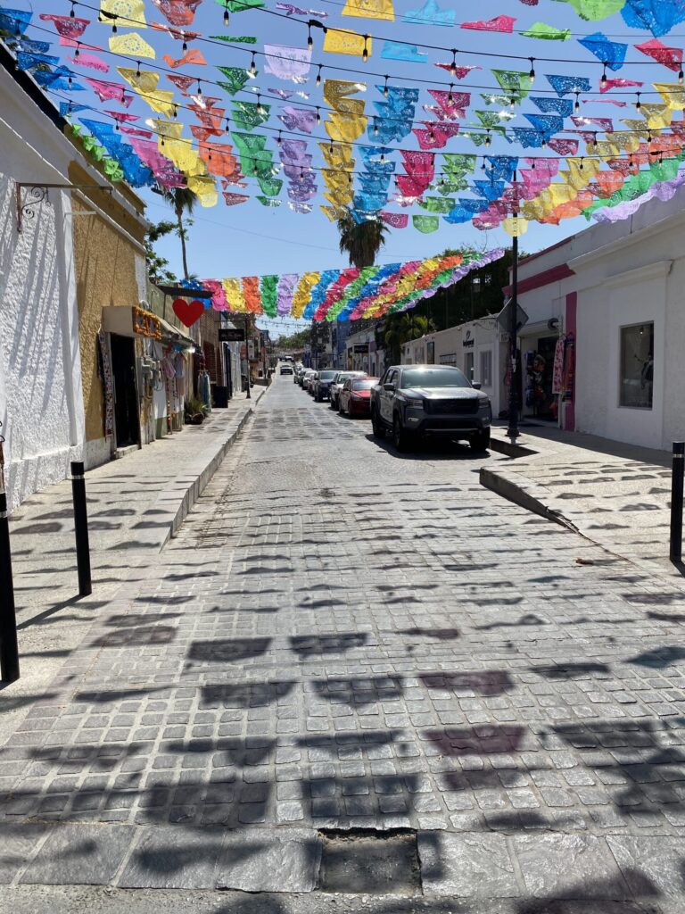 Sunny cobblestone street lined with white buildings and parked cars, colorful papel picado banners strung overhead.