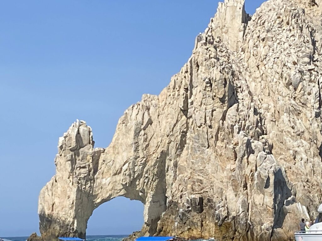 Rocky coastal arch jutting into the blue sea under a clear sky; people at the base for scale.