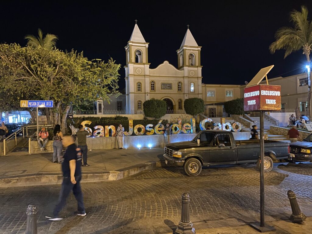Night view of a church with twin steeples in a courtyard, palm trees to the sides, and people gathered near a taxi stand.