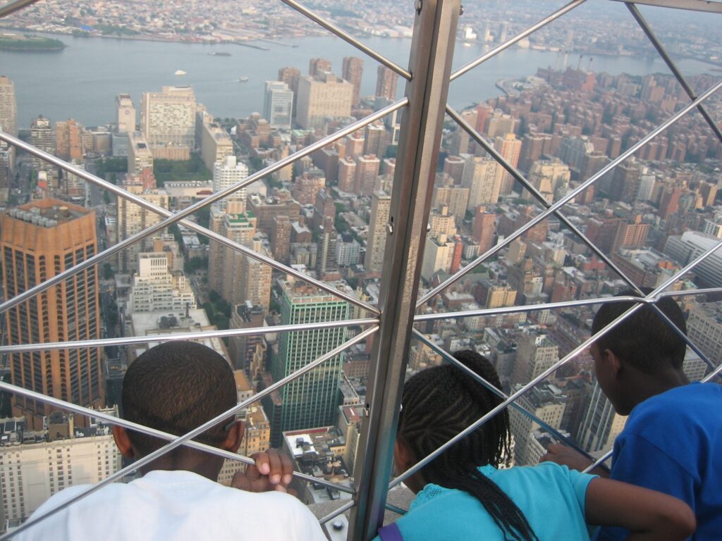 Three people lean against a glass railing on a high observation deck, overlooking a dense city with a river in the background.