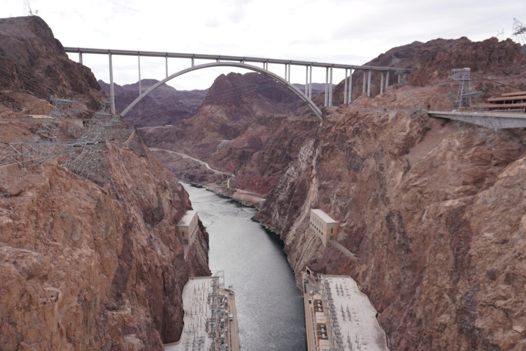 View of Hoover Dam with a large arch bridge spanning a deep canyon of red rock and rugged cliffs on both sides (aerial perspective).