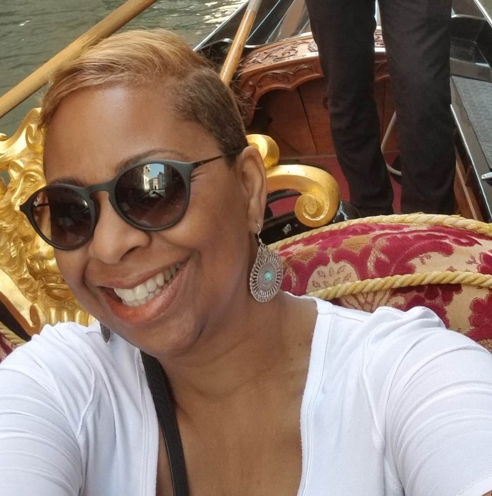 Smiling woman with short blond hair and sunglasses takes a selfie on a gilded, ornate boat with red upholstered seating behind her.