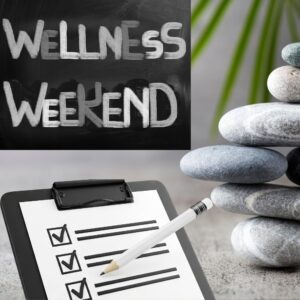 Chalkboard reads 'Wellness Weekend' beside a balanced stack of smooth stones and a clipboard checklist with a pencil.